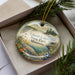 A decorative ornament in a box, featuring a scenic landscape with a lake, mountains, and pine trees.