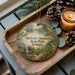A wooden tray with a decorative ornament featuring a scenic landscape of Georgia State Park, accompanied by pine cones and a lit candle.