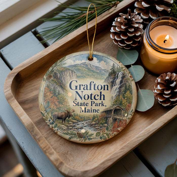 A decorative ornament featuring a scenic mountain landscape with a moose, hanging on a wooden tray next to pine cones and a lit candle.