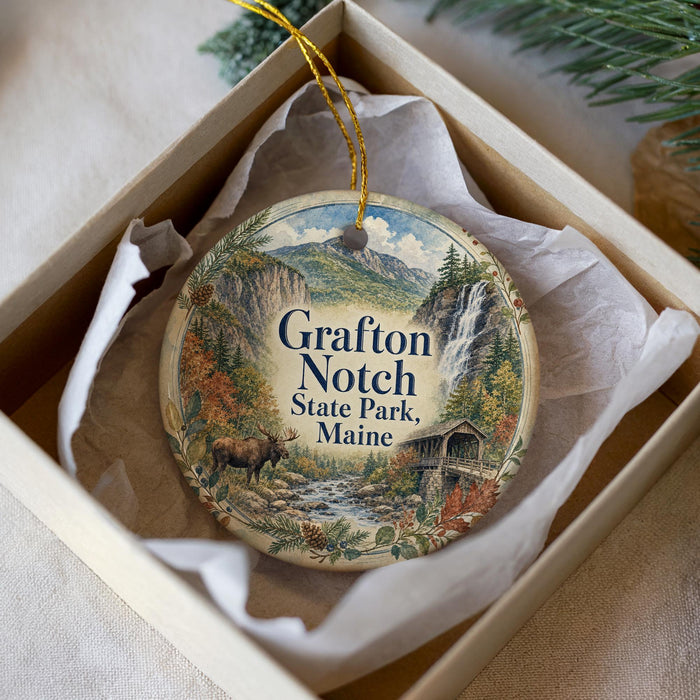 A decorative ornament in a box, featuring a scenic landscape with a waterfall, pine trees, and a moose.