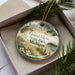 A decorative ornament in a box, featuring a scenic landscape with a lighthouse and pine trees, along with the text "Grand Mere State Park, Michigan".