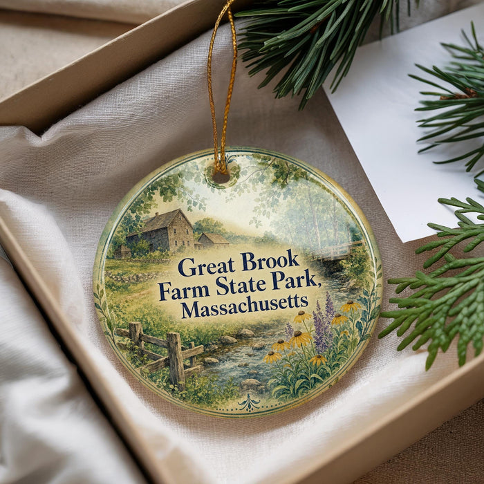 A round ornament with a scenic countryside image, featuring a farm, a stream, and pine trees, is displayed in a box with pine branches.