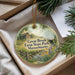 A round ornament with a scenic countryside image, featuring a farm, a stream, and pine trees, is displayed in a box with pine branches.