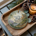 A decorative ornament featuring a scenic image of the Bluffs State Park in Minnesota, with pine cones and a lit candle on a wooden tray.