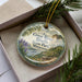 A decorative ornament in a box, featuring a scenic landscape with a river, mountains, and pine trees.