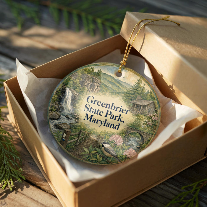 A circular ornament in a brown box, featuring a scenic landscape with a waterfall, trees, and a house, with the text "Greenbrier State Park, Maryland" printed on it.