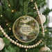 A round ornament hanging on a Christmas tree, featuring a scenic landscape with a river, trees, and a house.