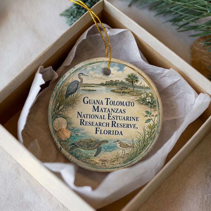 A round ornament with a beach scene and text, placed in a box with a white tissue paper lining.