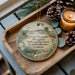 A decorative ornament with a bird and a landscape scene, placed on a wooden tray next to a lit candle and pine cones.