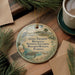 A round ornament with a beach scene and a bird is displayed on a wooden surface, accompanied by a cup of coffee and some papers.