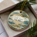 A decorative ornament in a box, featuring illustrations of a beach, pier, and birds, with the text "Gulf State Park, Alabama" prominently displayed.