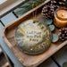 A wooden tray with a decorative ornament featuring a scenic landscape of a lake, trees, and birds, along with pine cones and a lit candle.