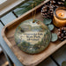 A wooden tray with a decorative ornament featuring a scenic image of Gunpolder Falls State Park, Maryland, along with pine cones and a lit candle.