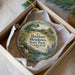 A decorative ornament in a box, featuring a scenic landscape with a house, trees, and a body of water.