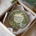 A round ornament with a scenic image of a park, featuring a bird, a bridge, and a river, is displayed in a box.
