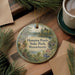 A round ornament with a scenic image of a pond and trees, surrounded by pine branches, sits on a wooden surface.
