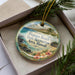 A decorative ornament with a tropical beach scene, featuring palm trees, flowers, and a body of water. The ornament is placed in a box with pine branches, creating a festive and holiday-themed display.