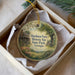 A round ornament with a painting of a farm and a house, placed in a box with a white tissue paper lining.