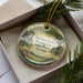 A decorative ornament in a box, featuring a scenic landscape with a house, trees, and flowers.