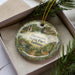 A decorative ornament in a box, featuring a scenic image of a park with a deer, a bridge, and a forest.