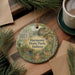 A decorative ornament with a scenic image of a park, featuring a bridge, trees, and a deer, hangs on a wooden surface alongside a cup of coffee and some pine branches.