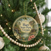 A round ornament hanging on a Christmas tree, featuring a scenic landscape with a waterfall and a cabin, surrounded by greenery and beaded ornaments.