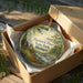 A round ornament in a brown box, featuring a scenic image of Hartwick Pines State Park, Michigan.