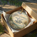 A round ornament in a box, featuring a beach scene with a pier and palm trees, along with the text "Henderson Beach State Park, Florida".