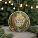 A circular ornament with a painting of a forest scene, featuring a waterfall and trees, hangs in front of a blurred background with Christmas lights.