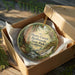 A round ornament in a brown box, featuring an illustration of a forest scene with a waterfall, placed on a wooden surface.