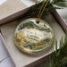 A decorative ornament in a box, featuring a scenic landscape with a deer, mountains, and a river.