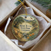 A round ornament with a scenic landscape and a deer, placed in a box with a white tissue paper lining.