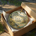 A round ornament in a wooden box, featuring a scenic landscape with a deer, mountains, and a river.