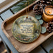 A decorative ornament with a house and trees, placed on a wooden tray next to a lit candle and pine cones.