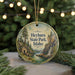 A round ornament with a scenic image of a mountain landscape, featuring a waterfall and pine trees, hangs in front of a blurred background with Christmas lights.