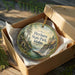 A decorative ornament in a wooden box, featuring a scenic landscape with a waterfall and pine trees.