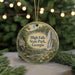 A round ornament with a painting of a waterfall and a house, surrounded by greenery, is displayed in front of a blurred background with Christmas lights.