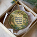 A decorative ornament in a white box, featuring a scenic illustration of a tropical landscape with palm trees, a bird, and a body of water.