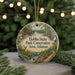 A round ornament with a scenic landscape featuring a river, forest, and a cabin, hanging in front of a Christmas tree with lights.