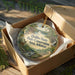 A round ornament in a box, featuring a scenic landscape with a river, trees, and a cabin, with the text "Hobbs State Park-Conservation Area, Arkansas" printed on it.