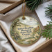 A decorative ornament with a painting of a house and trees, hanging from a string, placed inside a box with pine branches.
