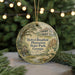 A circular ornament with a painting of a house and trees, surrounded by greenery, hangs in front of a blurred background with twinkling lights.