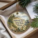 A round ornament with a lighthouse and a bird, hanging from a string, placed inside a box with pine branches and a white card.