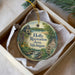 A round ornament with a scenic illustration of a river, trees, and a house, hanging from a string.