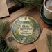 A round ornament with a scenic landscape of a river, trees, and a castle, surrounded by pine branches and a cup of coffee on a wooden surface.