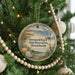 A round ornament hanging on a Christmas tree, featuring a beach scene with a lighthouse and birds.