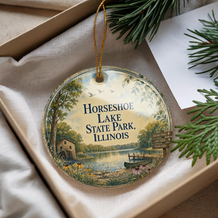 A decorative ornament with a scenic lake and forest landscape, featuring a house and a boat, is displayed in a box with pine branches.