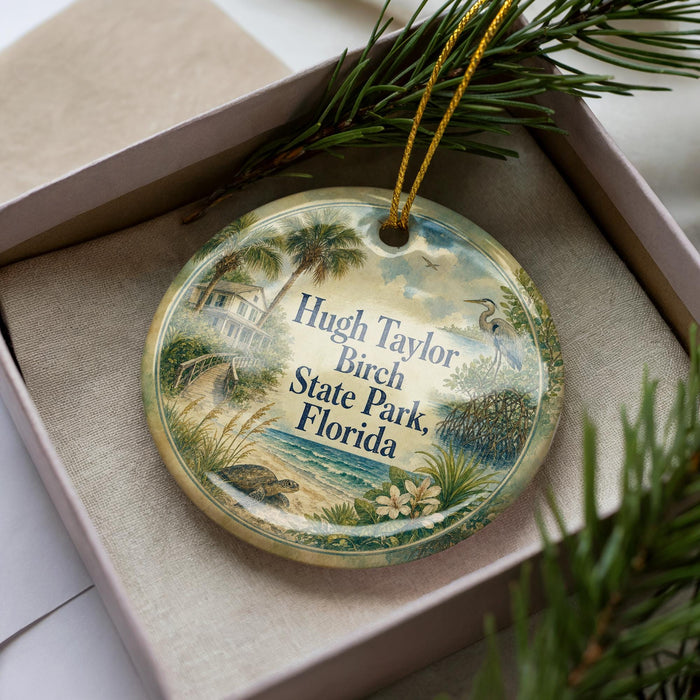 A decorative ornament with a tropical scene, featuring palm trees, a bird, and a body of water, is displayed in a box with pine branches.
