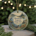 A round ornament with a scenic landscape featuring a lake, trees, and wildlife, including a crane, is displayed in front of a blurred background with Christmas lights.