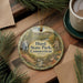 A decorative ornament with a scenic image of Hurd State Park in Connecticut, featuring a waterfall, a bridge, and autumn foliage. The ornament is hanging from a string and is placed on a wooden surface, surrounded by pine branches and a cup of coffee.
